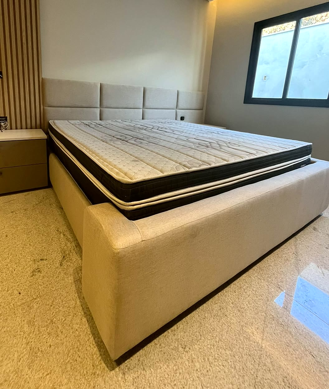 Cama estofada em linho bege com cabeceira de placas, em quarto com iluminação indireta